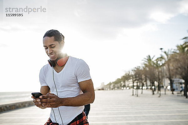 Spanien  lächelnder junger Mann mit Kopfhörern an der Strandpromenade beim Blick aufs Handy