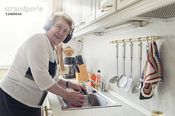 Porträt einer älteren Frau  die beim Spülen Musik mit Kopfhörern hört.