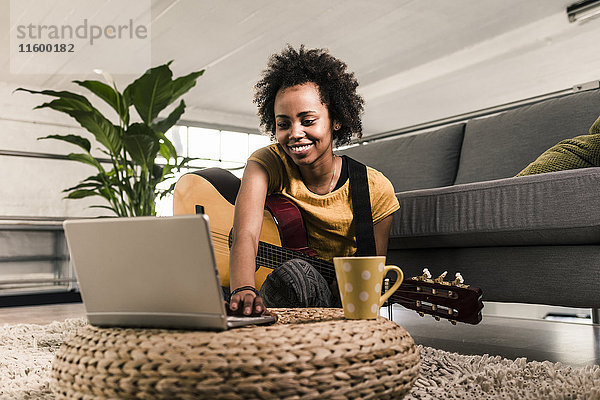 Lächelnde junge Frau zu Hause mit Gitarre und Laptop