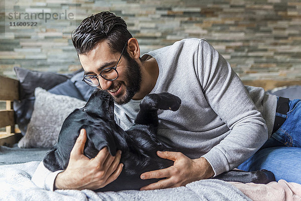 Lächelnder Mann  der mit seinem Hund auf der Couch zu Hause kuschelt.