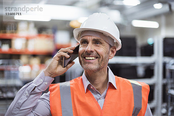 Lächelnder Mann in der Fabrikhalle mit Sicherheitsweste und Schutzhelm am Telefon