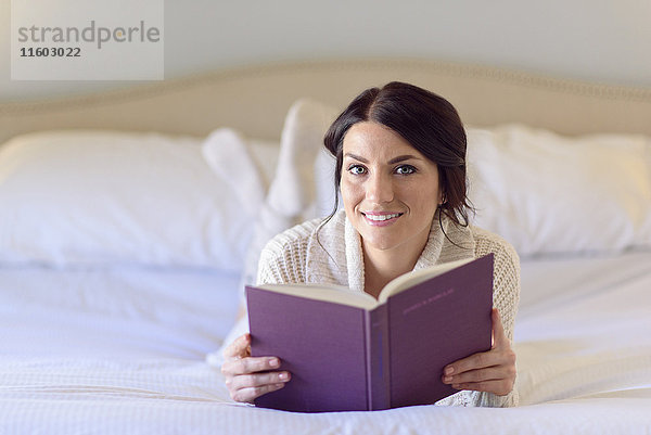 Lächelnde kaukasische Frau liegt auf dem Bett und liest ein Buch