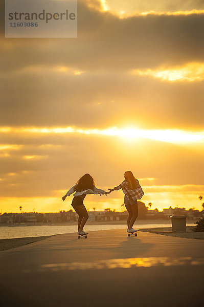 Mädchen hält sich an den Händen  während sie bei Sonnenuntergang mit dem Skateboard am Wasser fährt