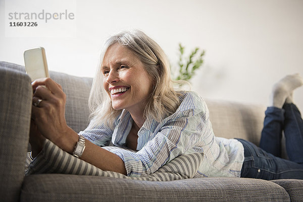 Lächelnde kaukasische Frau  die auf dem Sofa liegt und eine SMS auf ihrem Mobiltelefon schreibt
