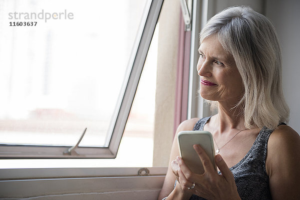 Lächelnde kaukasische Frau  die in der Nähe des Fensters eine SMS auf ihrem Handy schreibt
