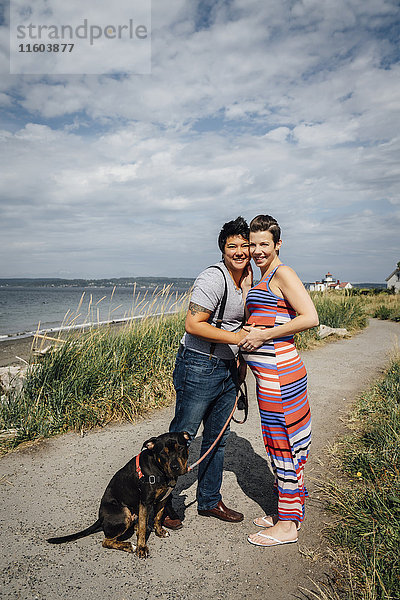 Porträt eines schwangeren lesbischen Paares mit Hund  das sich am Wasser umarmt