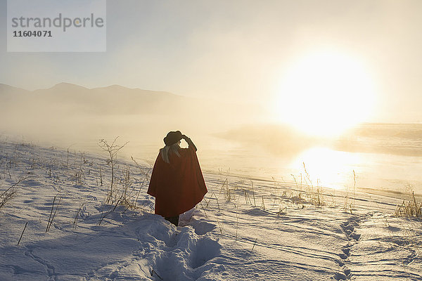 Kaukasische Frau  die ihre Augen in einer Winterlandschaft bei Sonnenuntergang abschirmt