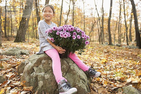 Lächelnde Mixed Race Mädchen sitzen auf einem Felsen im Herbst und halten Blumen