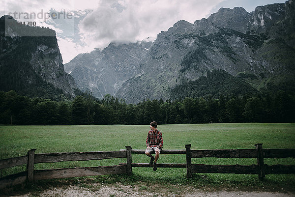 Kaukasischer Mann sitzt auf Holzzaun in der Nähe von Bergen