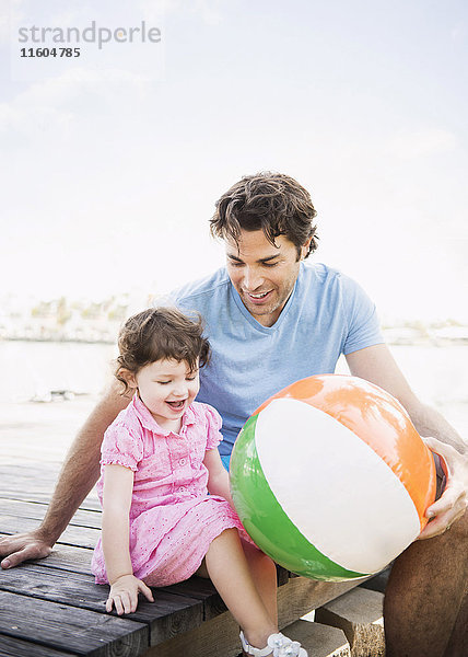 Kaukasischer Vater und kleine Tochter sitzen auf einem Steg mit Strandball