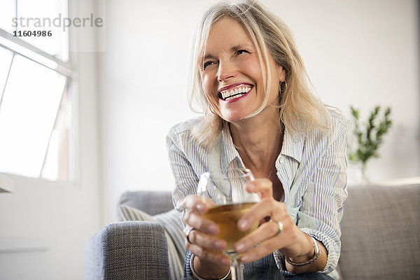 Lächelnde kaukasische Frau sitzt auf dem Sofa und trinkt Weißwein