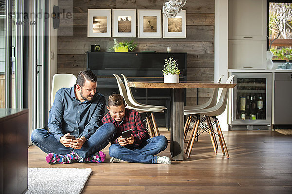 Lächelnder Vater und Sohn sitzen auf dem Boden und schreiben SMS auf ihren Handys