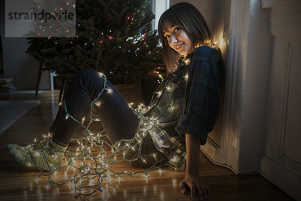 Mixed-Race-Frau sitzt auf dem Boden in Lichterketten eingewickelt in der Nähe von Weihnachtsbaum