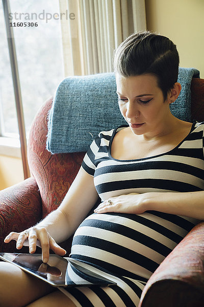 Schwangere kaukasische Frau  die in einem Sessel sitzt und ein digitales Tablet benutzt