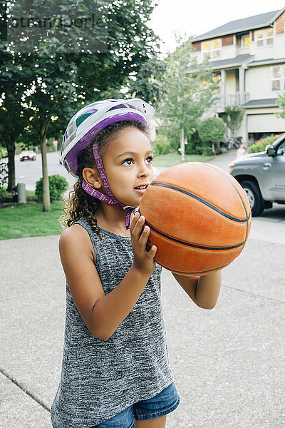 Gemischtrassiges Mädchen mit Helm spielt Basketball in der Auffahrt
