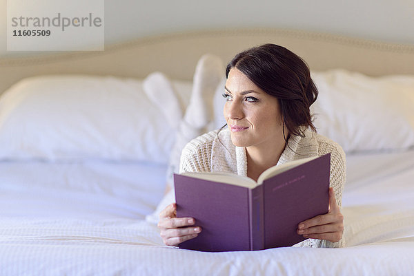 Nachdenkliche kaukasische Frau liegt auf dem Bett und liest ein Buch