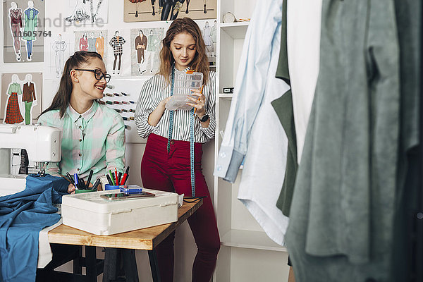 Fröhliche Designerin beim Blick auf weibliche Auszubildende mit Container im Designstudio