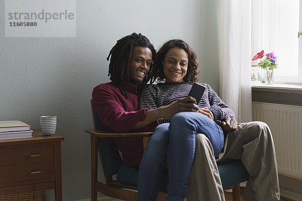 Lächelndes multiethnisches Paar  das zu Hause auf dem Sessel sitzt und ein Smartphone benutzt.