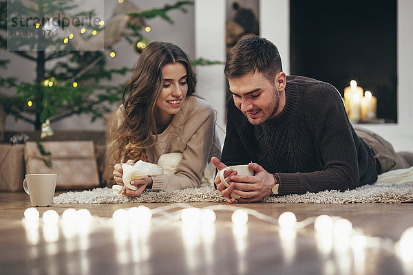 Lächelndes Paar beim Kaffeetrinken auf dem Teppich zu Hause zu Weihnachten