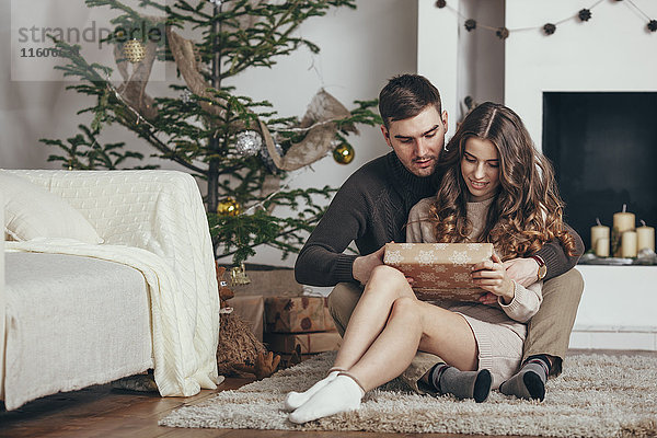 Junges Paar  das zu Weihnachten auf einem Teppich zu Hause sitzt und sich eine Geschenkbox ansieht.