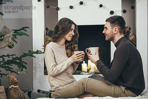 Lächelndes Paar beim Kaffeetrinken auf dem Teppich zu Hause zu Weihnachten
