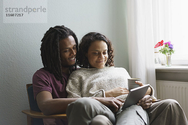 Junges multiethnisches Paar  das zu Hause auf einem Sessel sitzt und ein digitales Tablett benutzt.