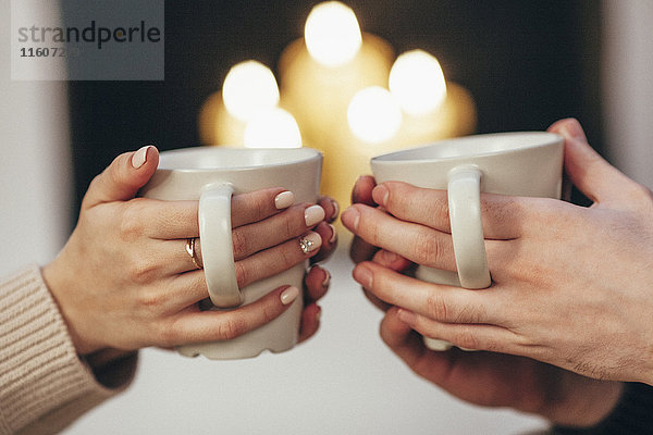 Beschnittenes Bild eines Paares  das Kaffeetassen gegen beleuchtete Kerzen zu Hause hält.