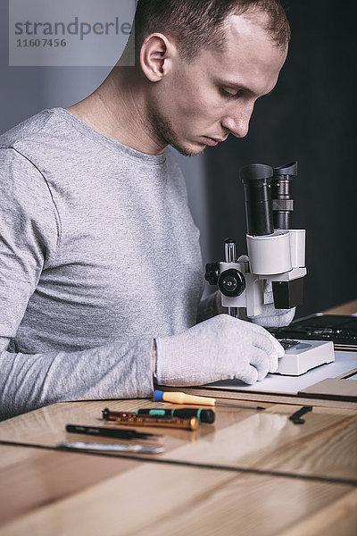 Junger Techniker untersucht Leiterplatte mit Mikroskop im Elektronikgeschäft