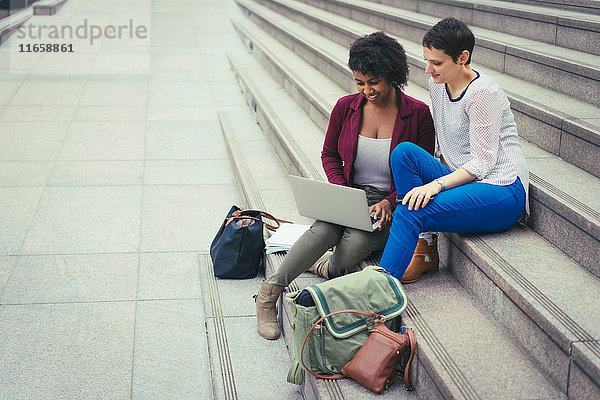 Zwei Frauen  die auf Stufen sitzen und auf einen Laptop schauen
