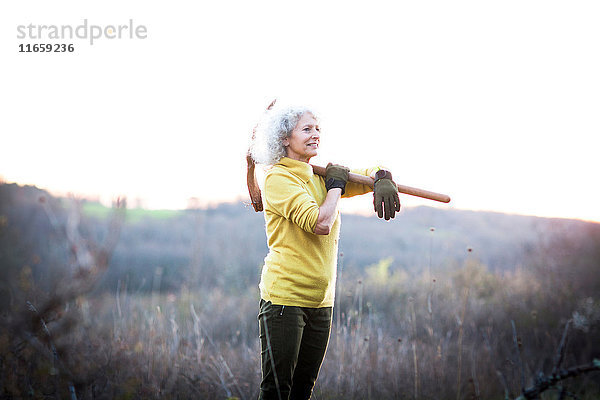 Reife Frau trägt in der Landschaft eine Spitzhacke über der Schulter