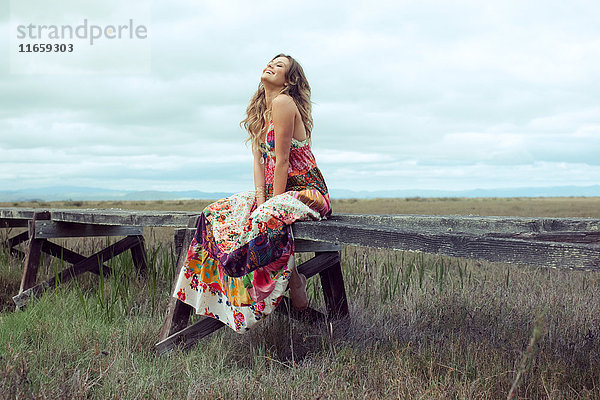 Junge Frau in Boho-Maxi-Kleid auf erhöhtem Holzsteg in Landschaft sitzend