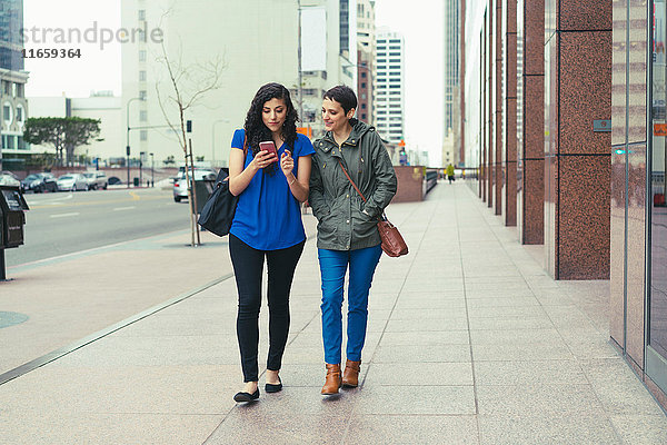 Zwei Freundinnen gehen die Straße entlang und schauen auf ein Smartphone