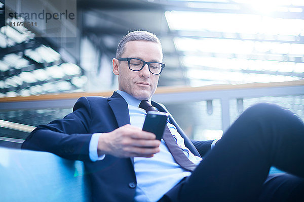 Geschäftsmann auf dem Sofa mit Blick auf Smartphone im Atrium des Büros