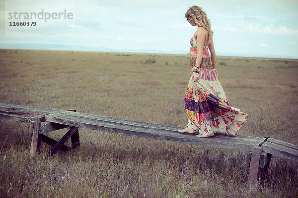 Junge Frau in Boho-Maxi-Kleid geht auf einem erhöhten Holzsteg in der Landschaft