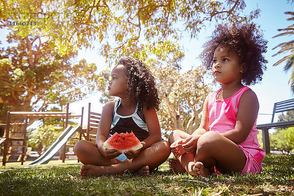 Zwei junge Schwestern  die auf Gras sitzen und Wassermelone essen