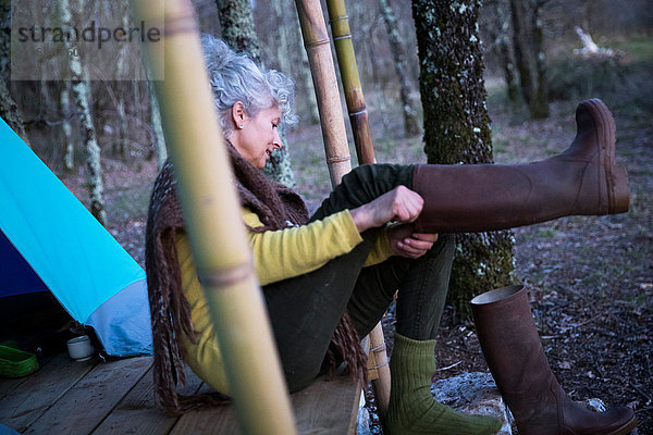 Reife Frau sitzt auf einer Waldveranda und zieht kniehohe Stiefel an