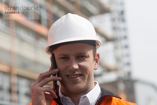 Porträt eines jungen männlichen Bauingenieurs  der auf einer Baustelle mit einem Smartphone telefoniert