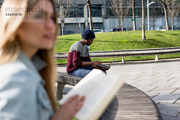 Junger Mann und Frau sitzen getrennt auf Bank  Mann mit Laptop  Frau hält Buch