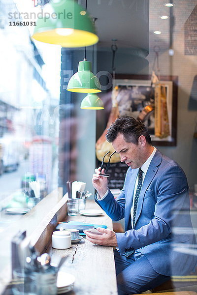 Der reife Geschäftsmann schaut auf das Smartphone im Restaurant-Fenstersitz