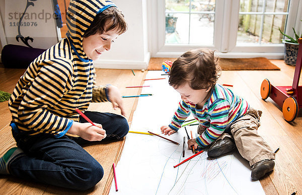 Männliches Kleinkind und großer Bruder sitzen auf dem Boden und zeichnen auf langem Papier