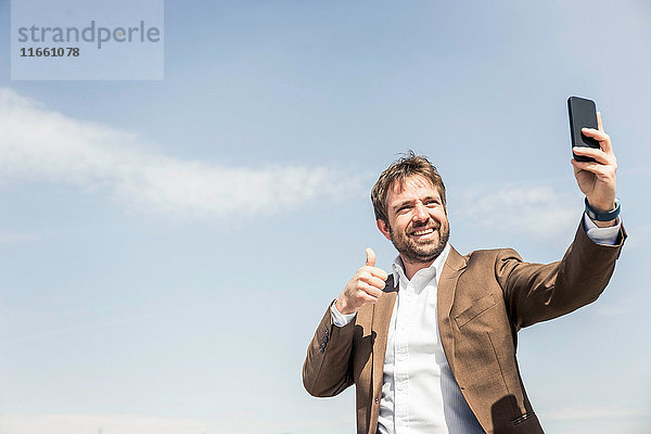 Geschäftsmann gibt Daumen hoch für Smartphone-Selfie gegen blauen Himmel