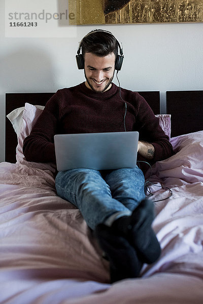 Junger Mann entspannt sich im Bett  benutzt Laptop  trägt Kopfhörer