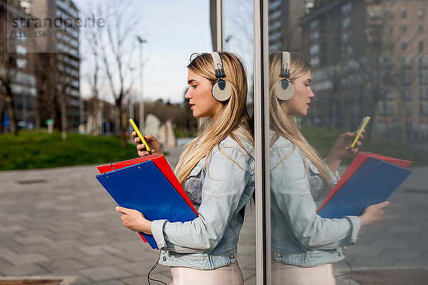 Junge Frau  im Freien  am Fenster lehnend  Kopfhörer tragend  Smartphone- und Dokumentendateien haltend