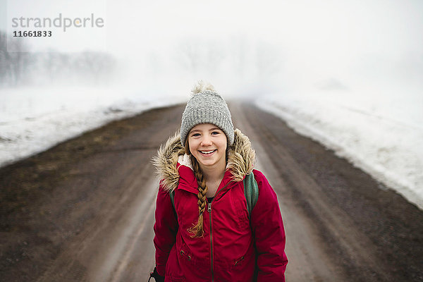 Porträt eines Mädchens mit Strickmütze  das mitten auf einem Feldweg im Nebel steht