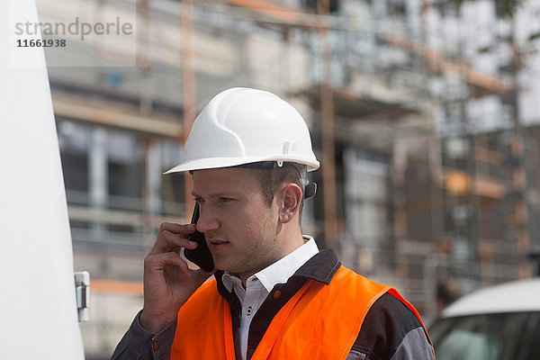 Junger männlicher Bauingenieur telefoniert mit Smartphone auf der Baustelle