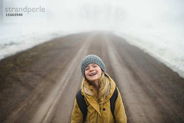 Porträt eines Mädchens mit Strickmütze  das lachend inmitten einer nebligen Schotterstraße steht