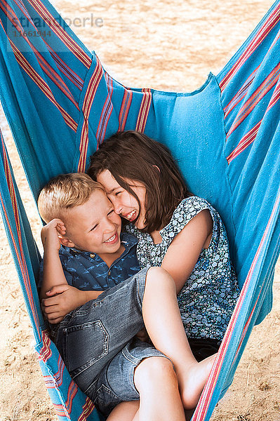 Mädchen und Bruder lachen zusammengekauert in der Hängematte am Poetto-Strand  Cagliari  Italien