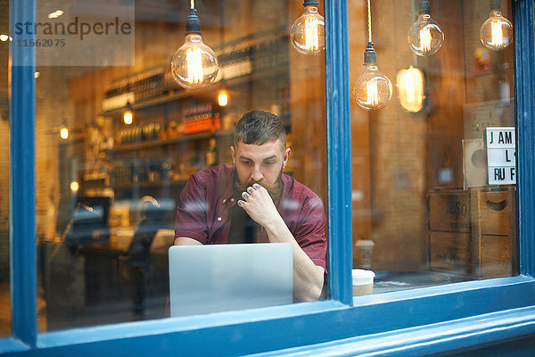 Fensteransicht des jungen Mannes mit Laptop im Cafe