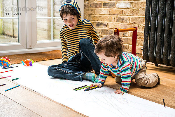 Männliches Kleinkind und großer Bruder sitzen auf dem Boden und zeichnen auf langem Papier