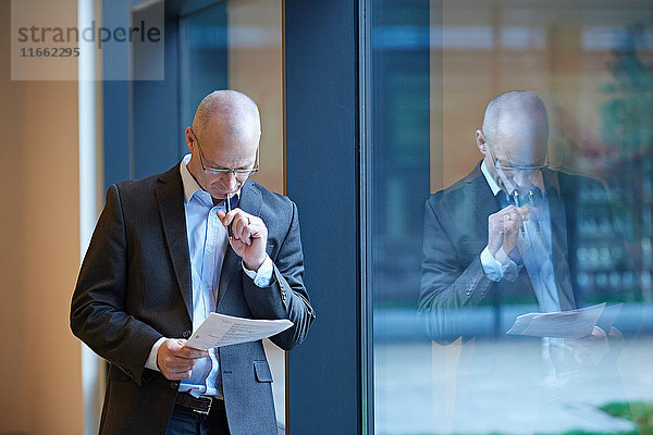 Reifer Geschäftsmann am Bürofenster beim Lesen von Papierkram